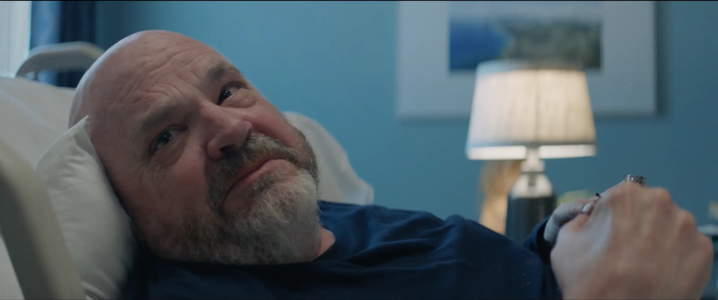 An older man rests in a hospital bed by a lamp.