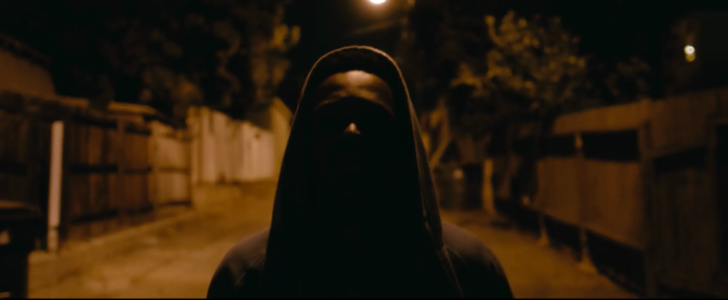 A hooded figure walks down a dark street at night.