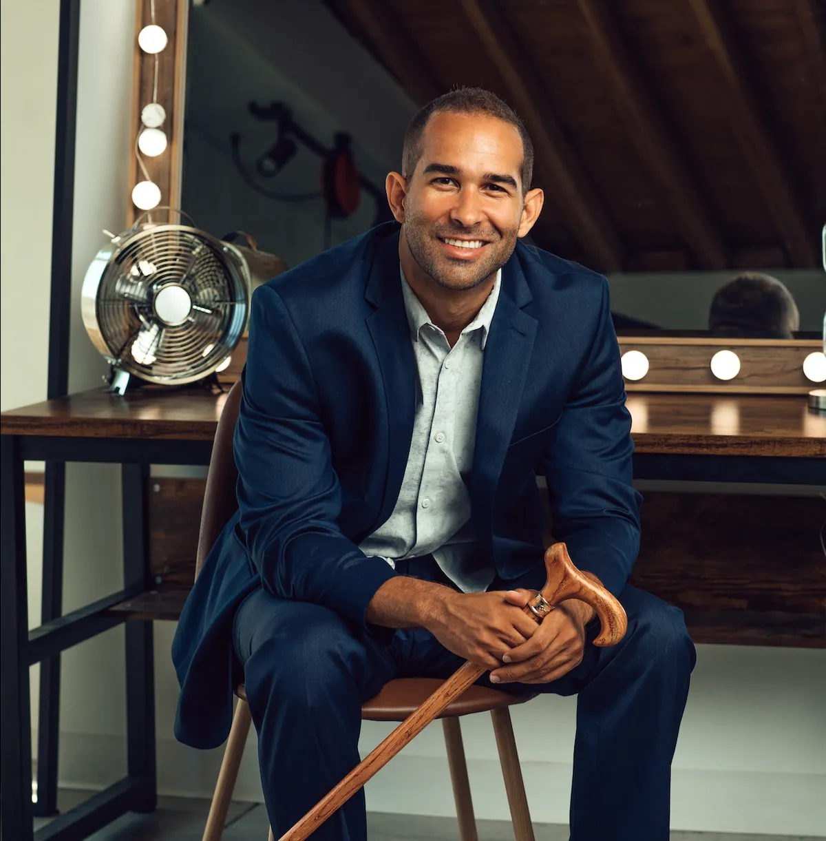 Portrait of Andrew Reid in a navy suit, seated with his cane in front of a lit dressing-room mirror.